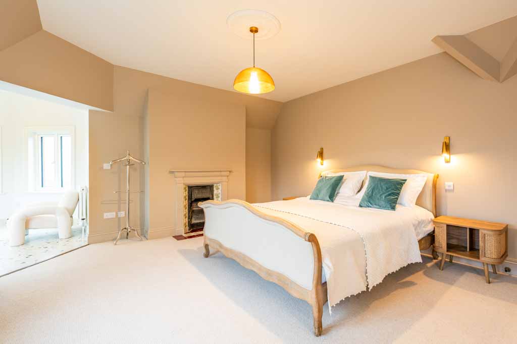 Bright and spacious bedroom featuring a wooden bed with white bedding and green accent pillows, two bedside tables, soft beige walls, and warm lighting from wall sconces and a ceiling lamp. The room has a calm and welcoming atmosphere, with a fireplace and an adjacent sitting area visible through an open doorway.