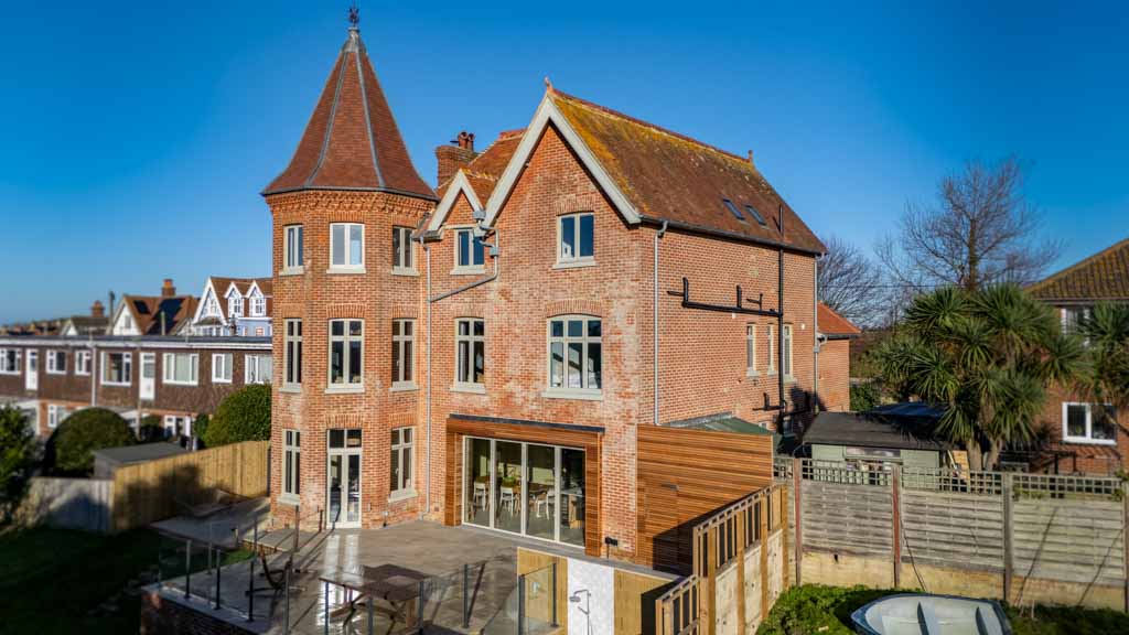 Large red brick house with a pointed turret and multiple tall windows, set in a residential neighbourhood with clear blue sky and neighbouring houses visible. The house has a modern glass extension and a spacious patio area, creating a welcoming and impressive atmosphere.